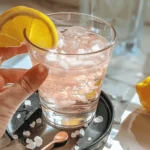 Hand holding a glass of pink salt diet drink with ice cubes and lemon slice garnish on a black coaster.