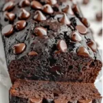A thick slice of moist chocolate zucchini bread with flecks of green, resting on a rustic wooden board with extra slices and a small bowl of sour cream in the background.