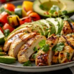 Close-up of grilled chicken breast topped with sliced avocado and lime.